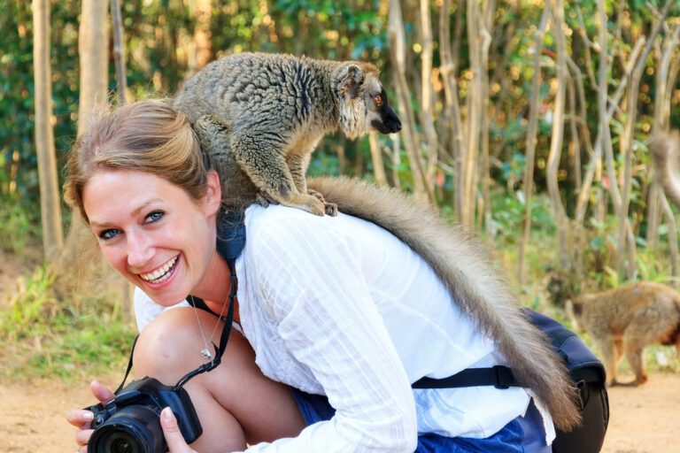 Reiziger gehurkt en lachend naar de camera met een camera in de handen en een lemur aap op haar rug