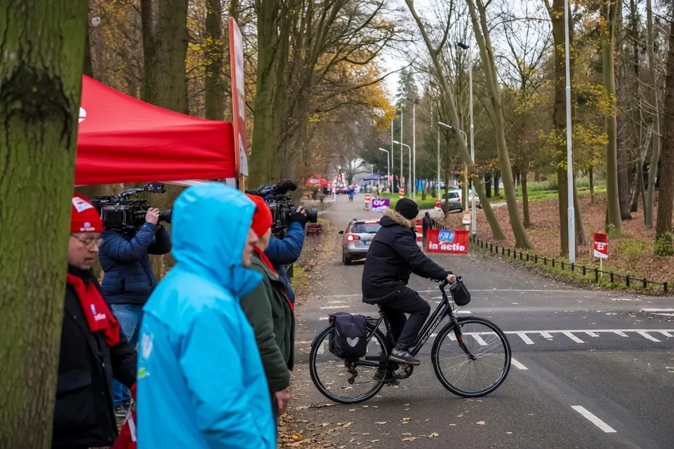 Mensen staan langs een weg bij een rood tentje, terwijl een fietser passeert