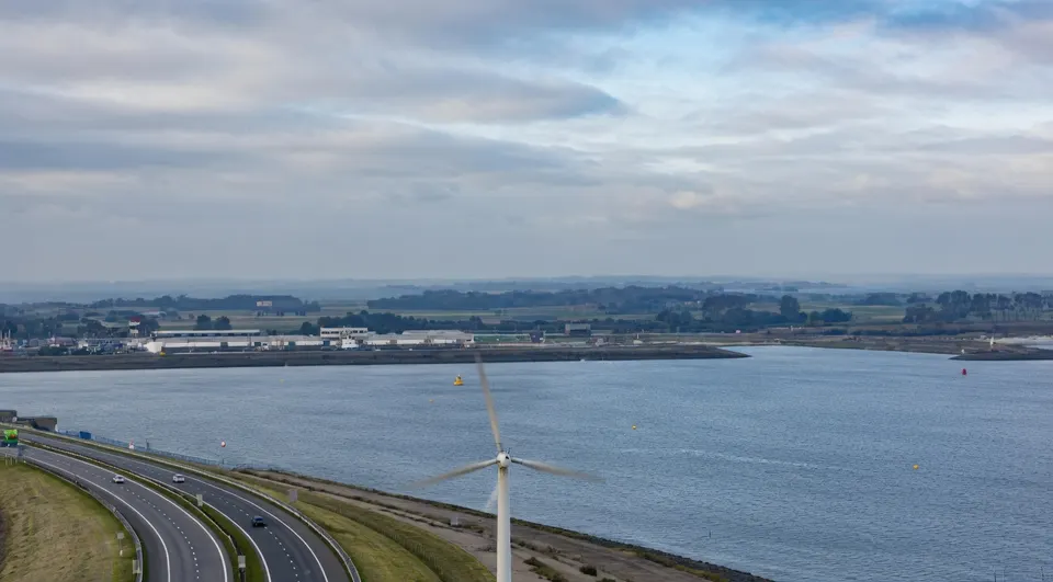 Windturbine langs een snelweg en water, met bebouwing en landbouwgrond op de achtergrond