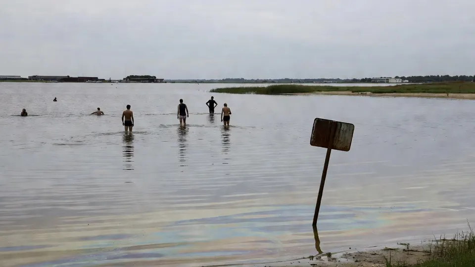 Zwemmers lopen door water bij een bord dat waarschuwt voor de kwaliteit van het zwemwater