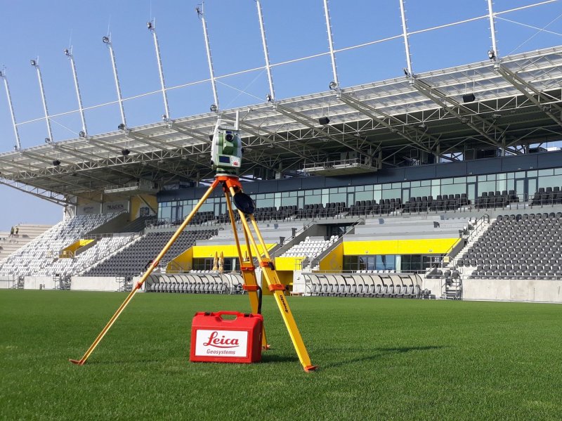 Leica Geosystems meracie zariadenie na statív na futbalovom ihrisku s tribúnami v pozadí.