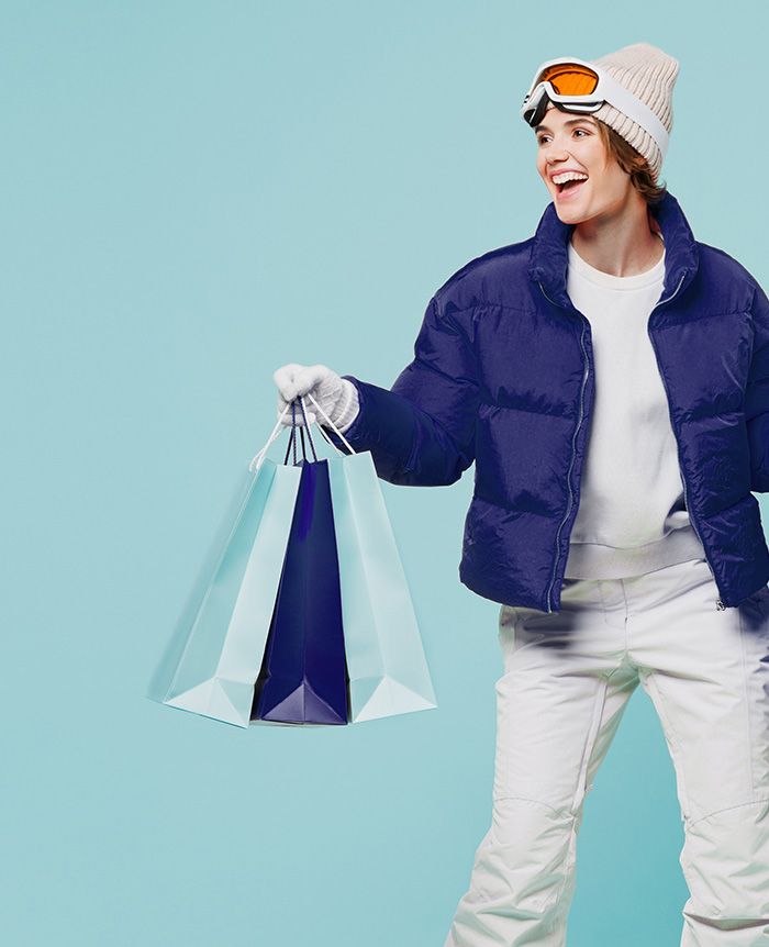 Person in weißer Mütze, oranger Skibrille, blauer Steppjacke und weißer Hose hält mehrere Einkaufstüten in der rechten Hand.