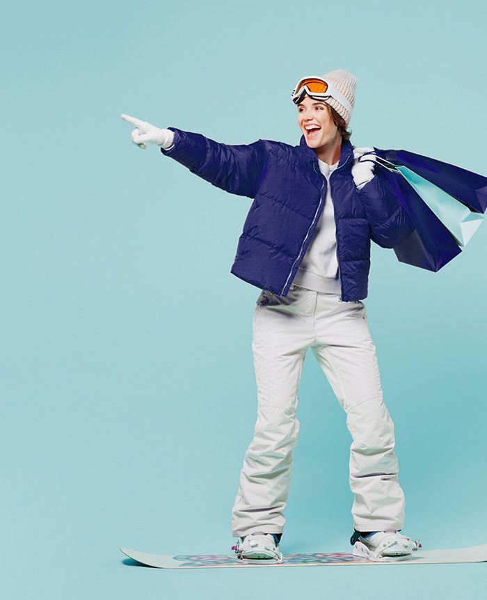 Person in weißer Skihose, blauer Jacke, weißer Mütze und Skibrille steht auf Snowboard und zeigt mit ausgestrecktem Arm nach links, trägt Einkaufstüten. 