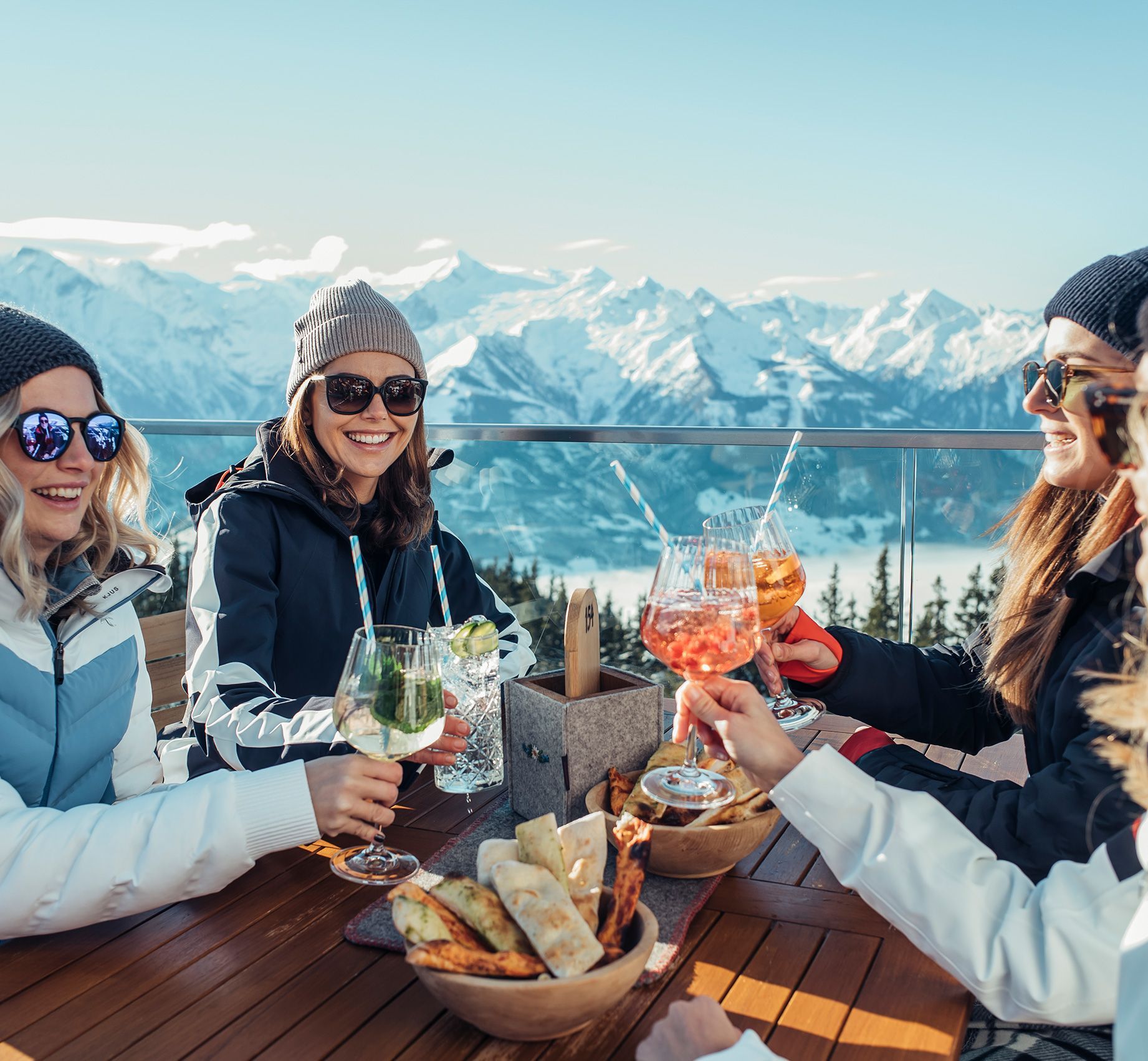 Vier Personen in Winterkleidung sitzen an einem Holztisch auf einer Terrasse mit Bergpanorama und stoßen mit Getränken in großen Gläsern an.
