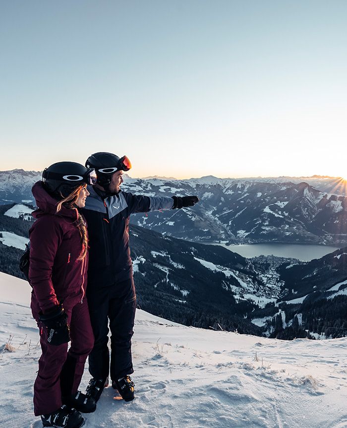 Zwei Skifahrer in Winterkleidung und Helmen stehen auf schneebedecktem Berg und blicken auf ein Tal mit See und Bergen im Hintergrund.