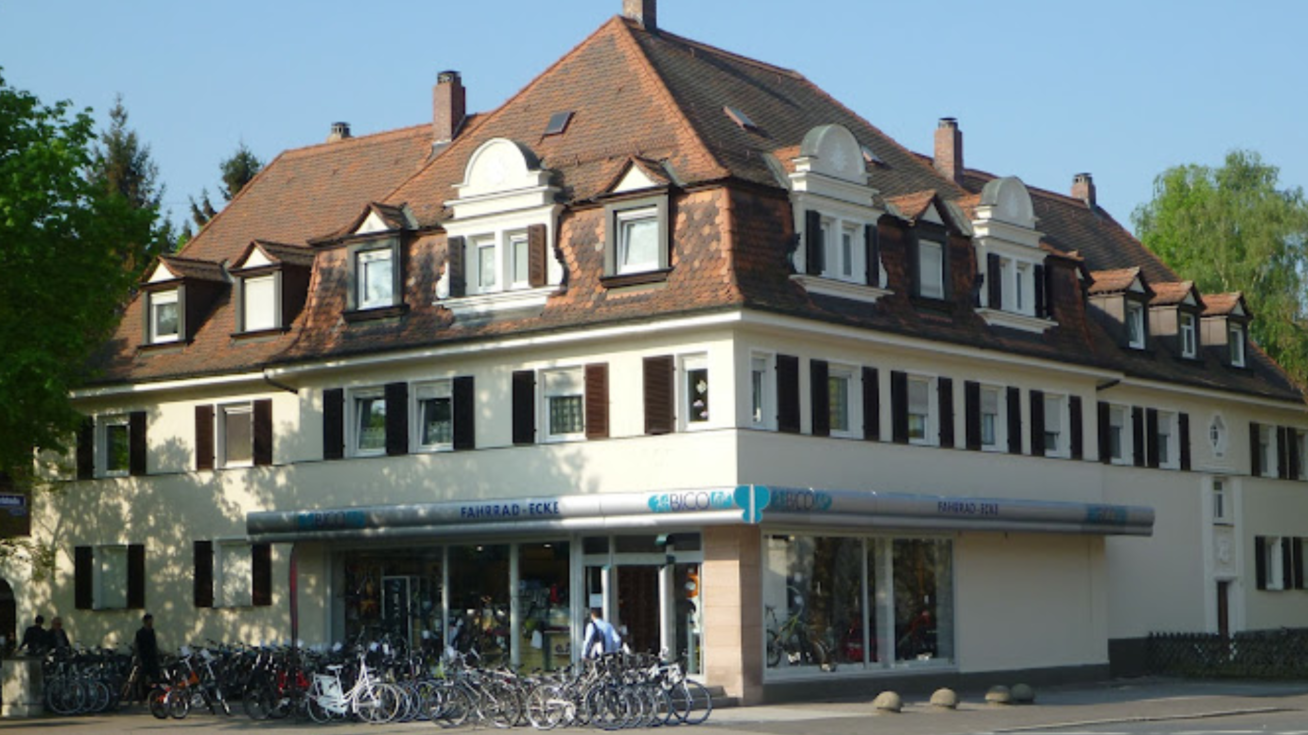 Bild eines historischen dreistöckigen Gebäudes mit Schindeldach und Dachfenstern. Im Erdgeschoss befinden sich Geschäfte mit Fahrrädern sowie eine Apotheke. Vor dem Gebäude parken zahlreiche Fahrräder.