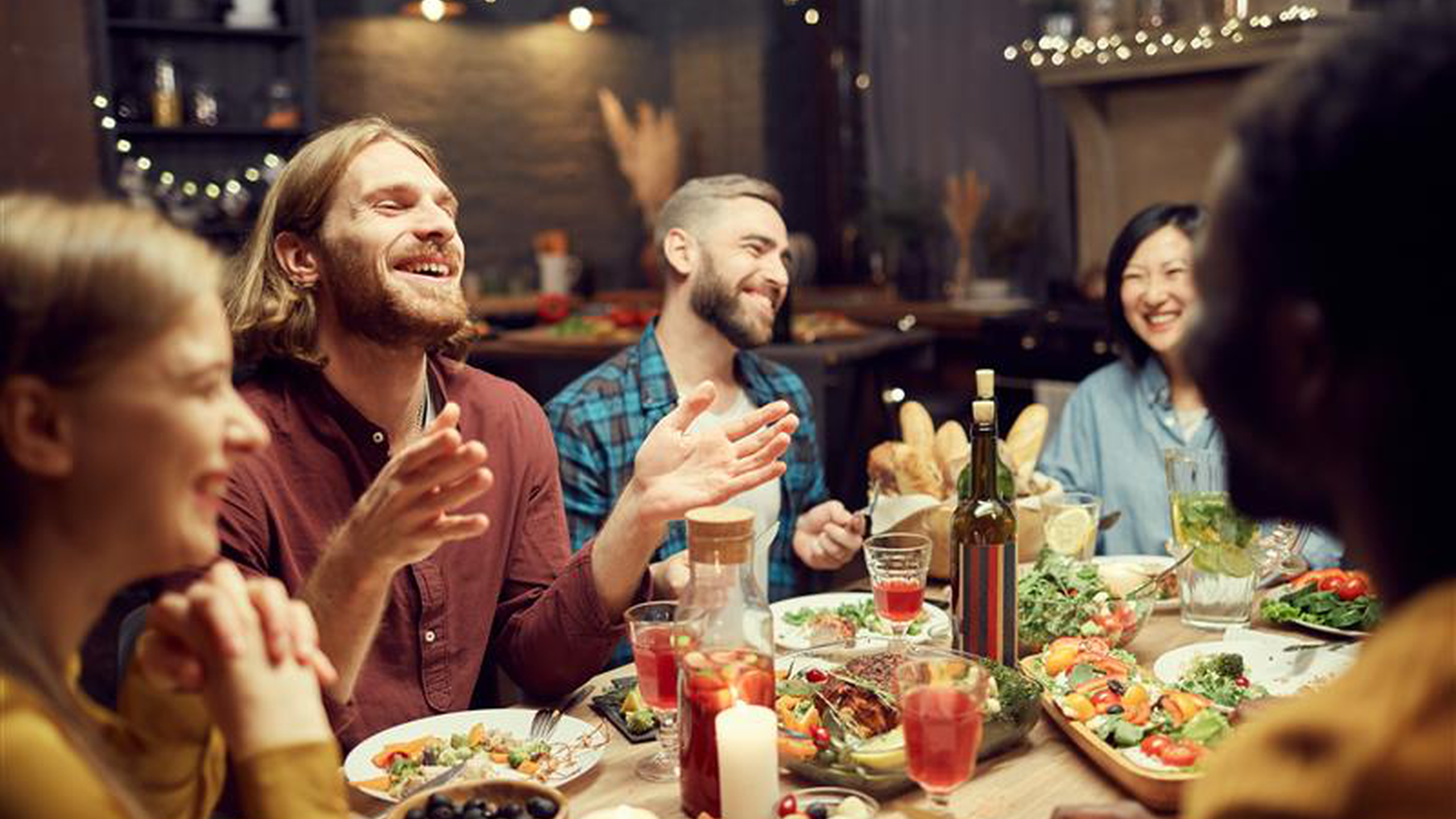 Fröhliche Freunde beim Abendessen um einen Tisch mit viel Essen und Getränken; sie unterhalten sich und lachen in einem gemütlichen Raum mit sanfter Beleuchtung.