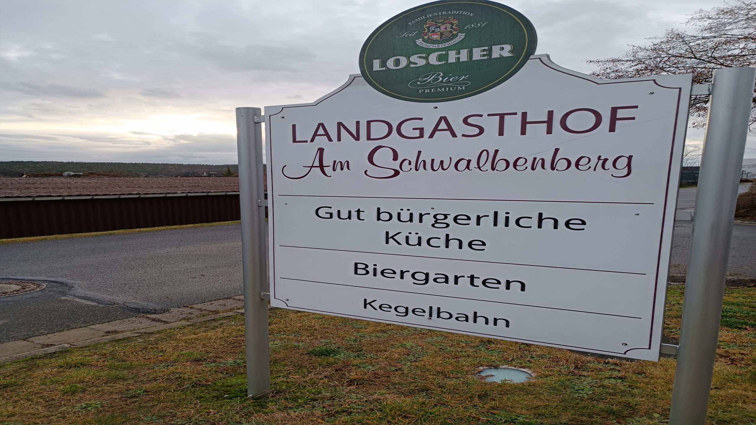 Wegweisendes Schild für den "Landgasthof Am Schwalbenberg" mit Angaben zu gut bürgerlicher Küche, Biergarten und Kegelbahn vor einem Hintergrund mit bewölktem Himmel.