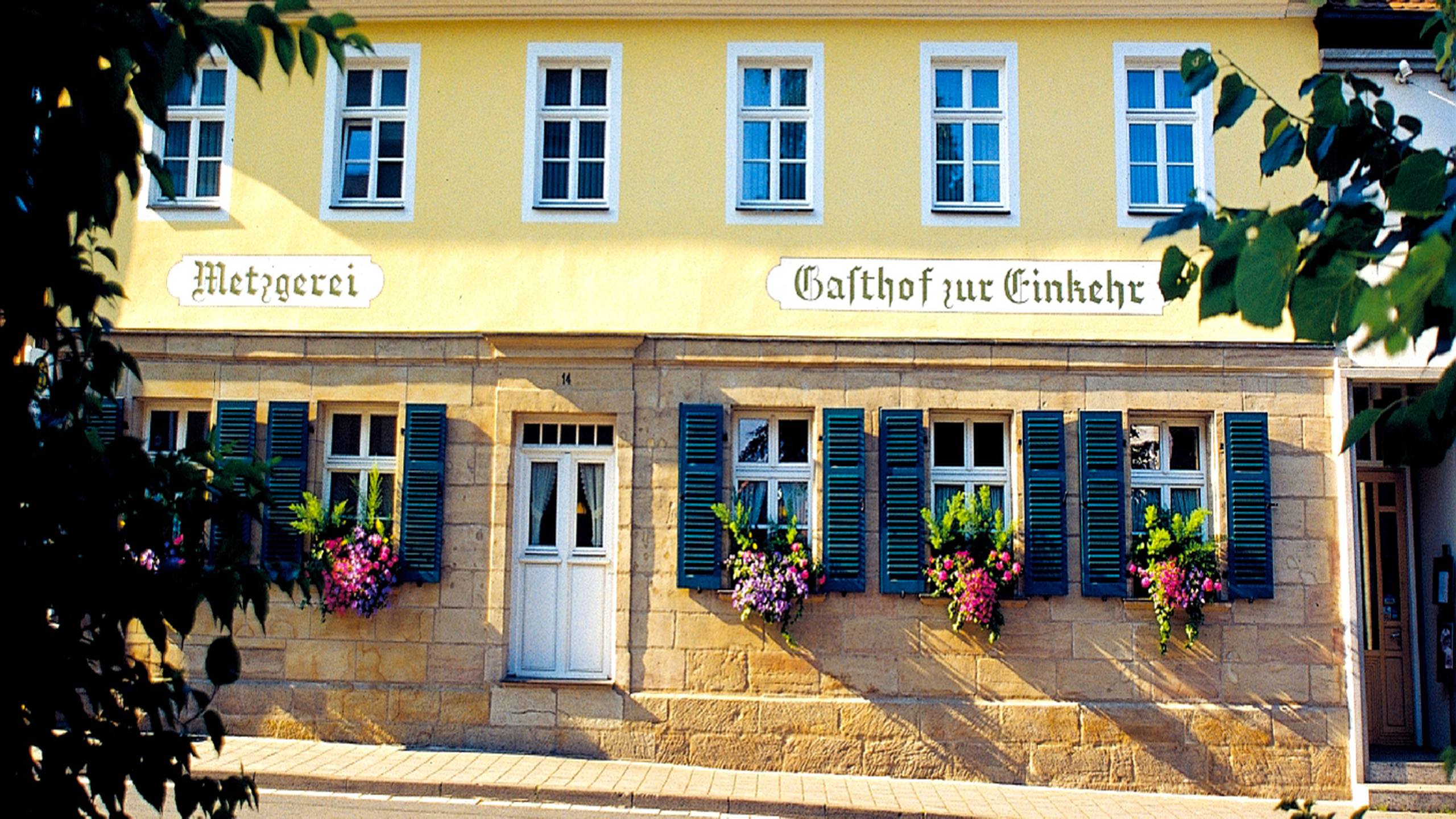 Historisches Gebäude in Gelb mit grünen Fensterläden, beschriftet als "Metzgerei" und "Gasthof zur Einkehr", geschmückt mit blühenden Blumenkästen.