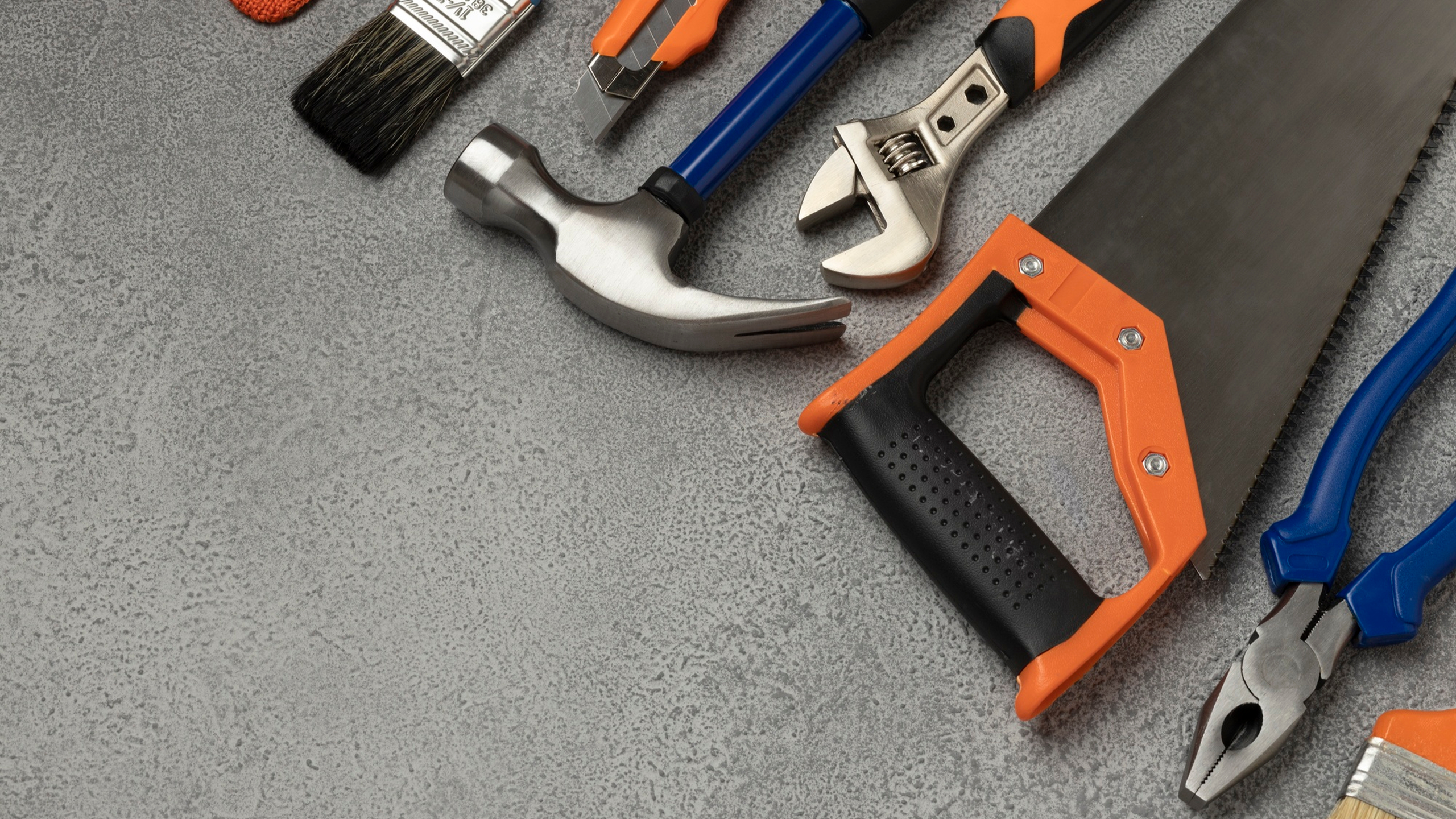 Werkzeuge auf grauer Oberfläche: Hammer, Pinsel, Schraubendreher, verstellbarer Schraubenschlüssel, Handsäge und Zangen verteilt.