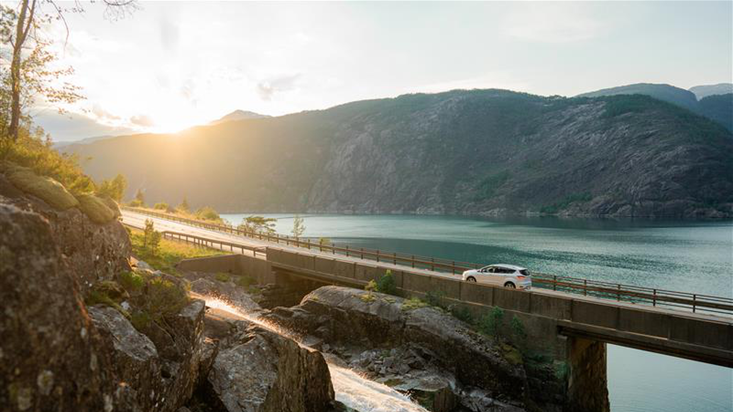Sonnenuntergang hinter Bergen, leuchtende das Auto auf einer Brücke entlang eines grünen Sees, umgeben von steilen Felswänden und einer bewaldeten Landschaft.