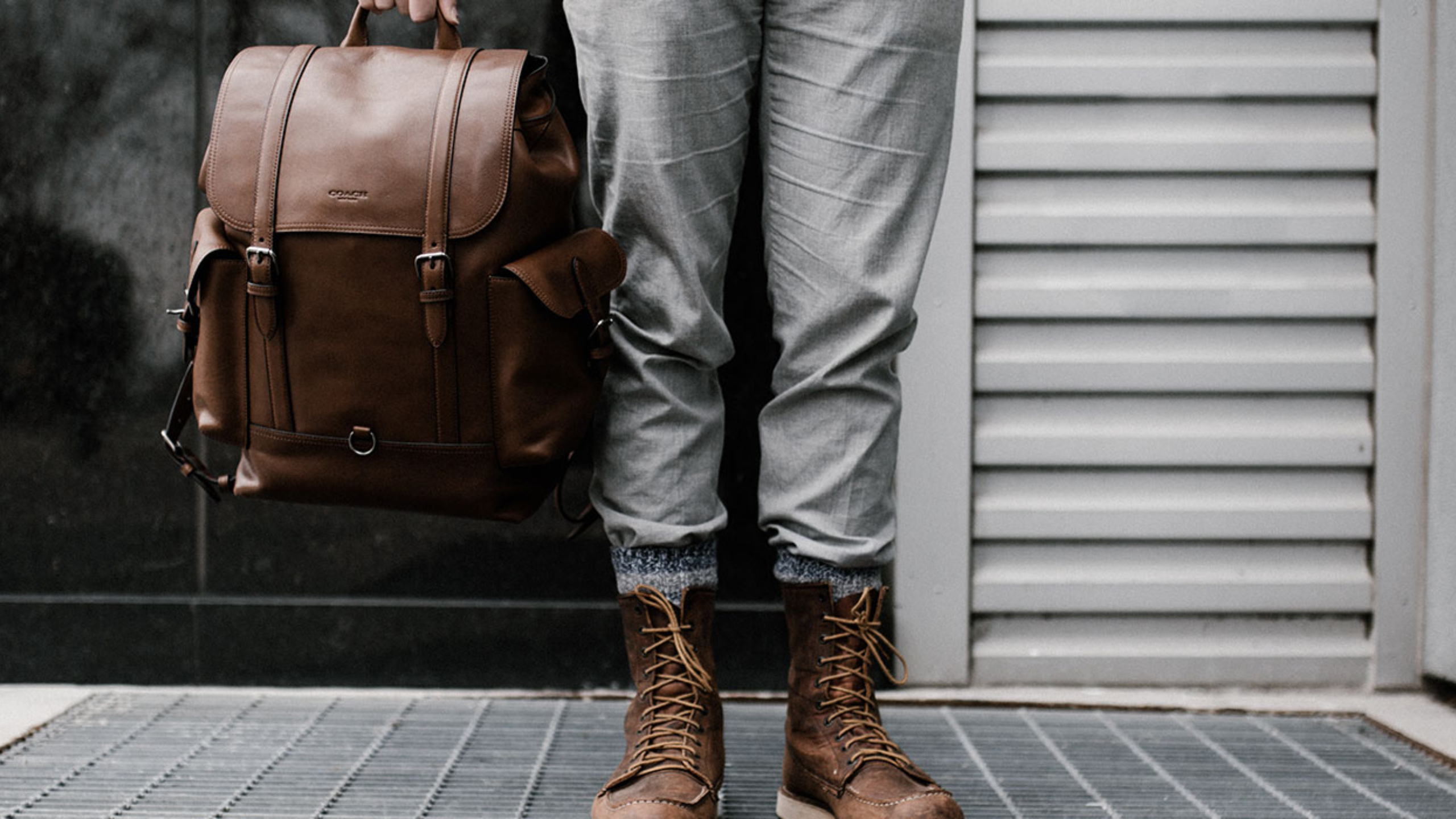 Eine Person steht vor einer grauen Wand und hält eine braune Ledertasche. Nur die untere Körperhälfte ist sichtbar; sie trägt graue Jeans und braune Schnürstiefel.