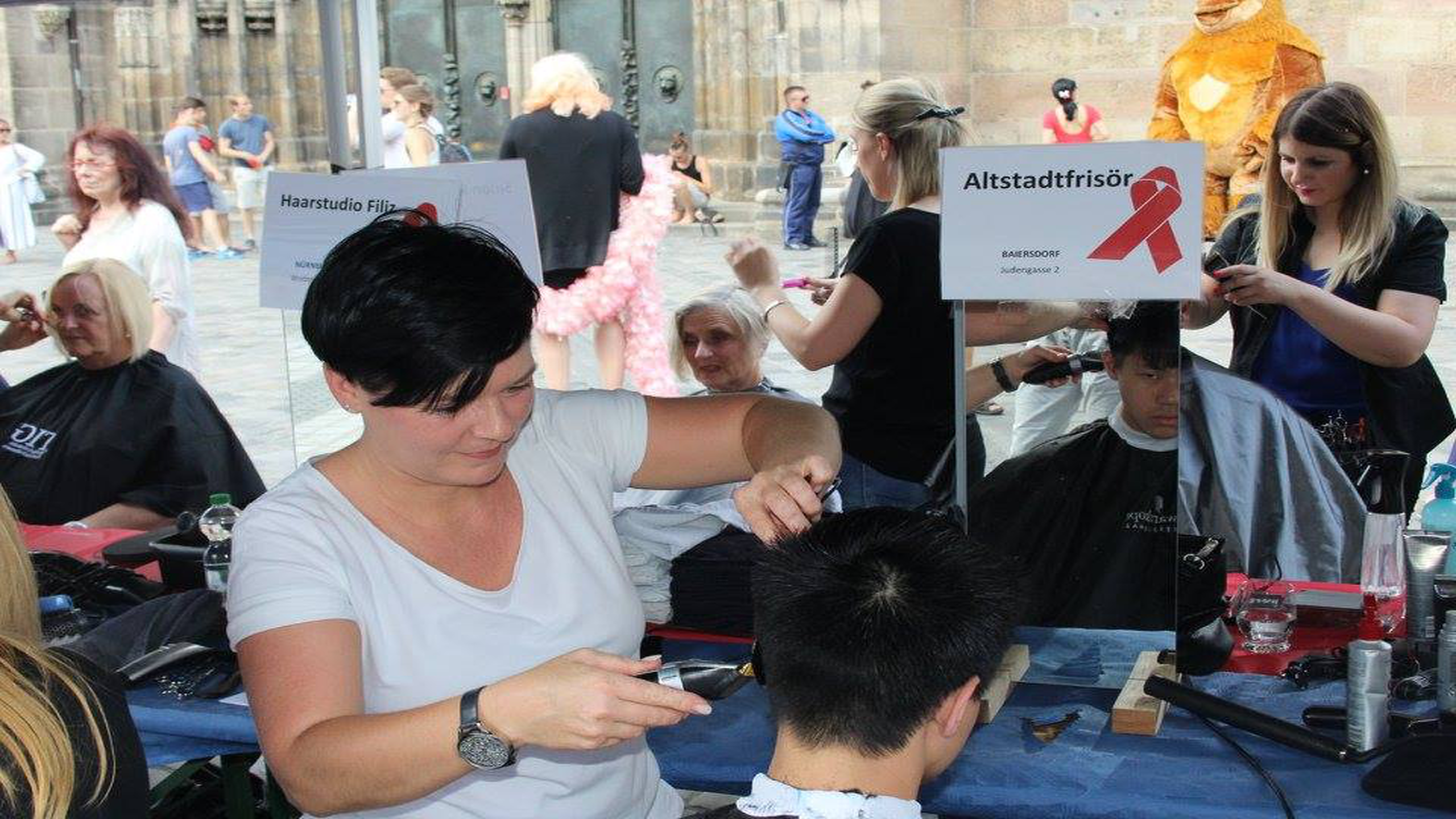 Friseurinnen schneiden Kunden im Freien die Haare bei einem öffentlichen Event. Schilder auf Tischen zeigen Salonnamen. Hintergrund zeigt Menschen und Stadtgebäude.