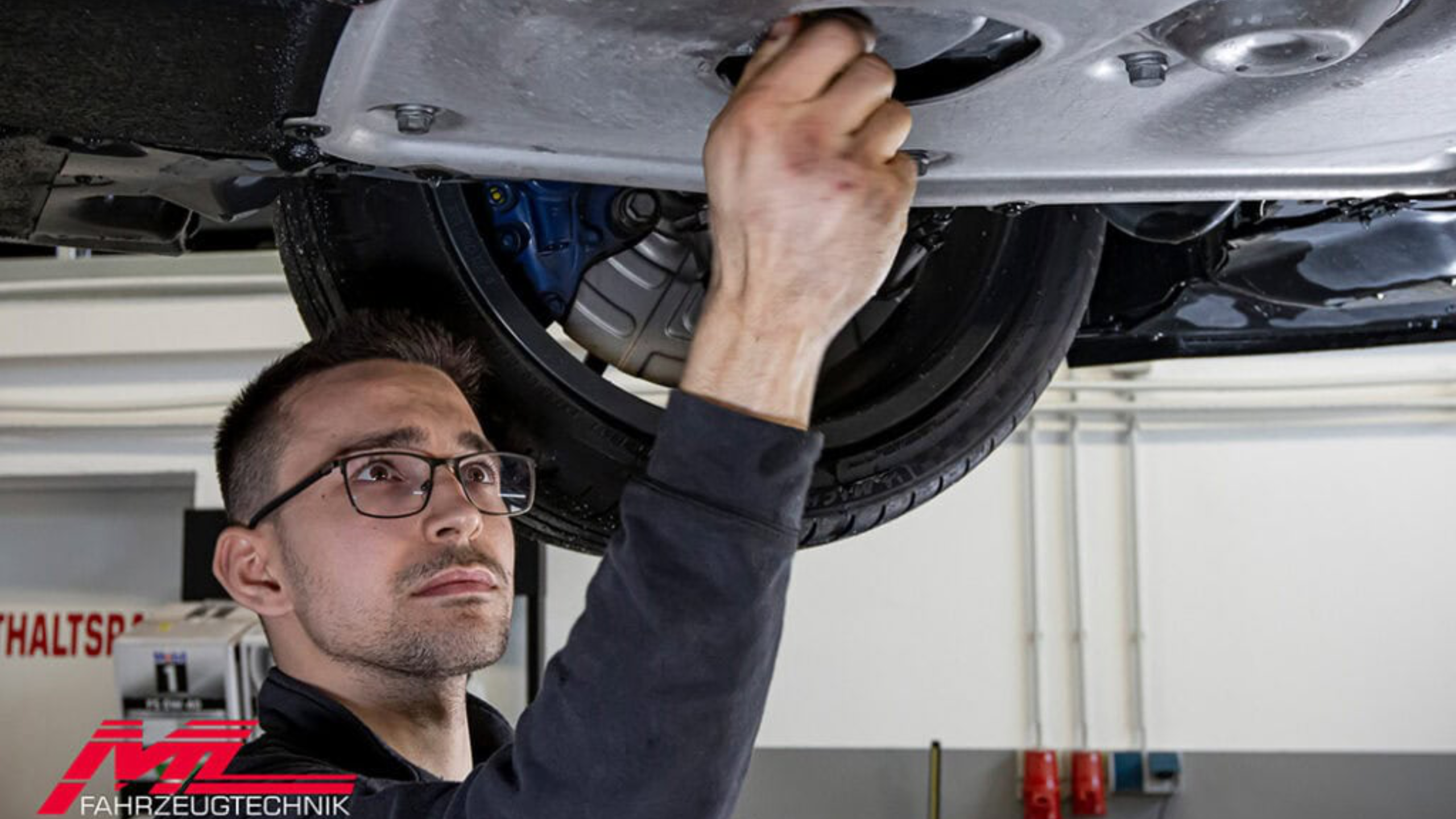 Ein Mechaniker mit Brille arbeitet fokussiert an der Unterseite eines Autos, welches auf einer Hebebühne ist. Er verwendet Werkzeug am Fahrwerk.