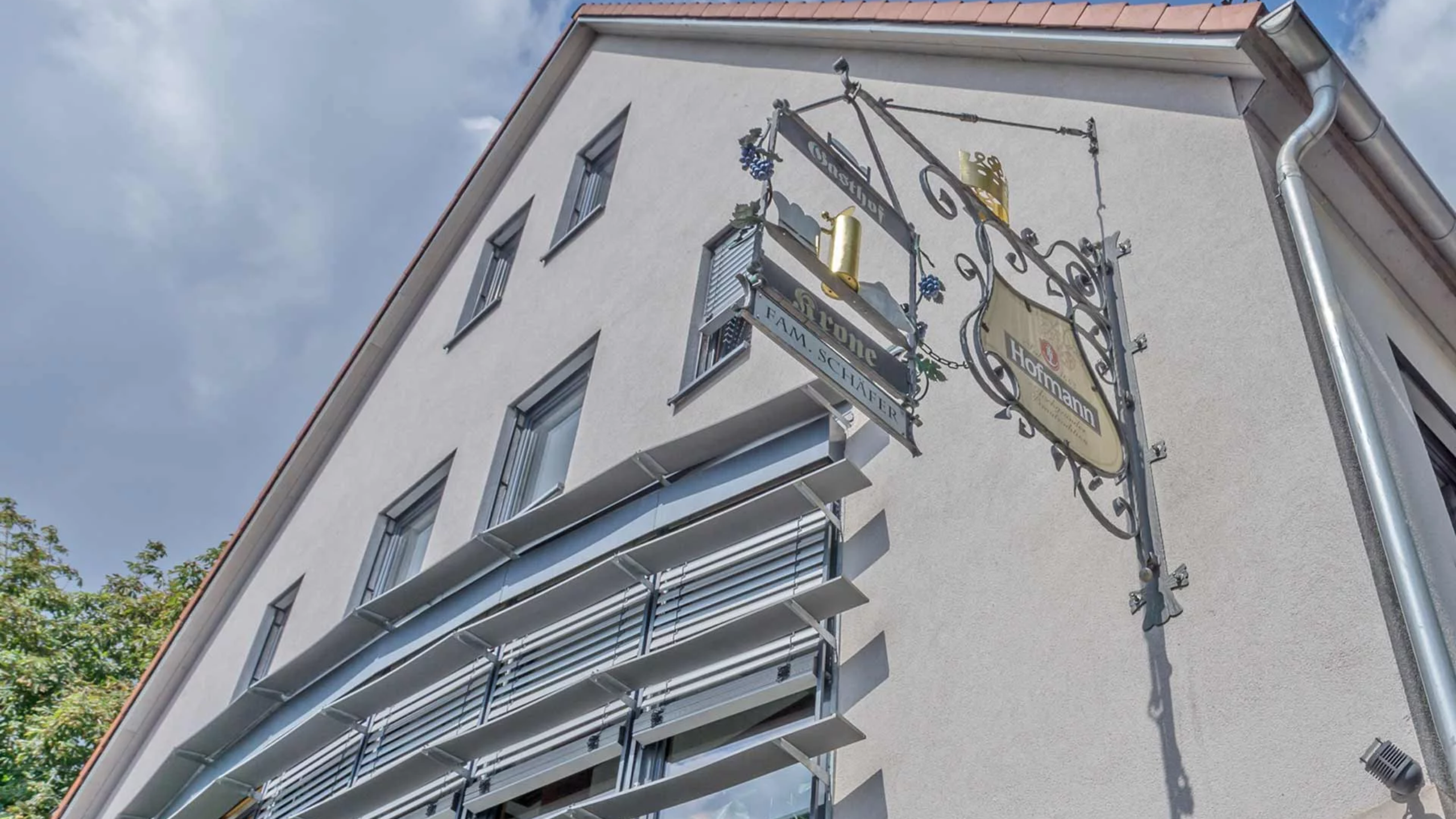 Weißes Gebäude mit grauen Jalousien, an dessen Ecke ein kunstvoll geschmiedetes Gasthausschild prangt, verziert mit Trauben und goldenem Weinbecher.