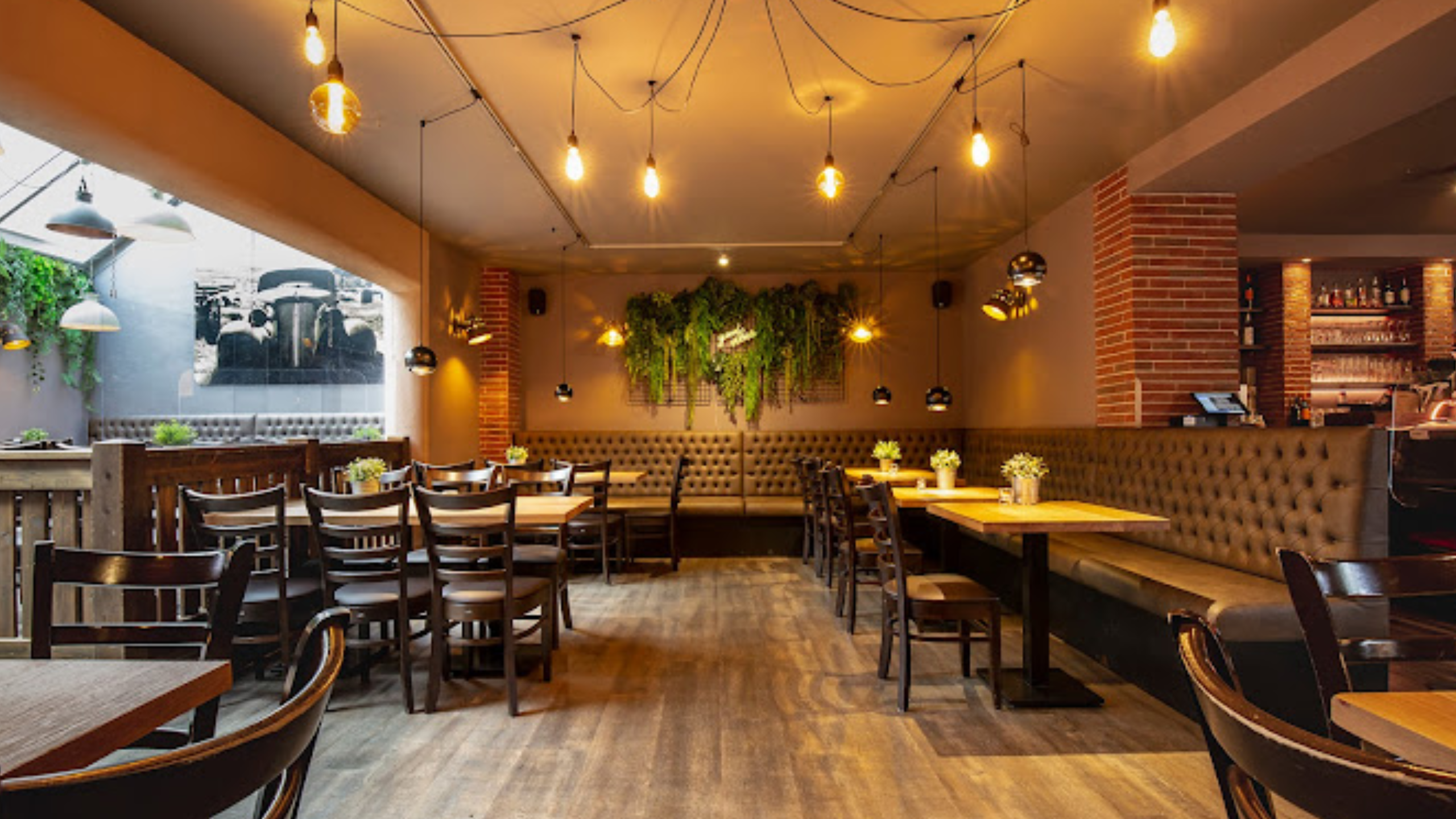 Gemütliches Restaurantinterieur mit Holzmöbeln, Sitzbänken und herabhängenden Glühbirnen. Wände sind mit Backstein und grünen Pflanzen dekoriert, oben rechts eine Bar mit Getränken.