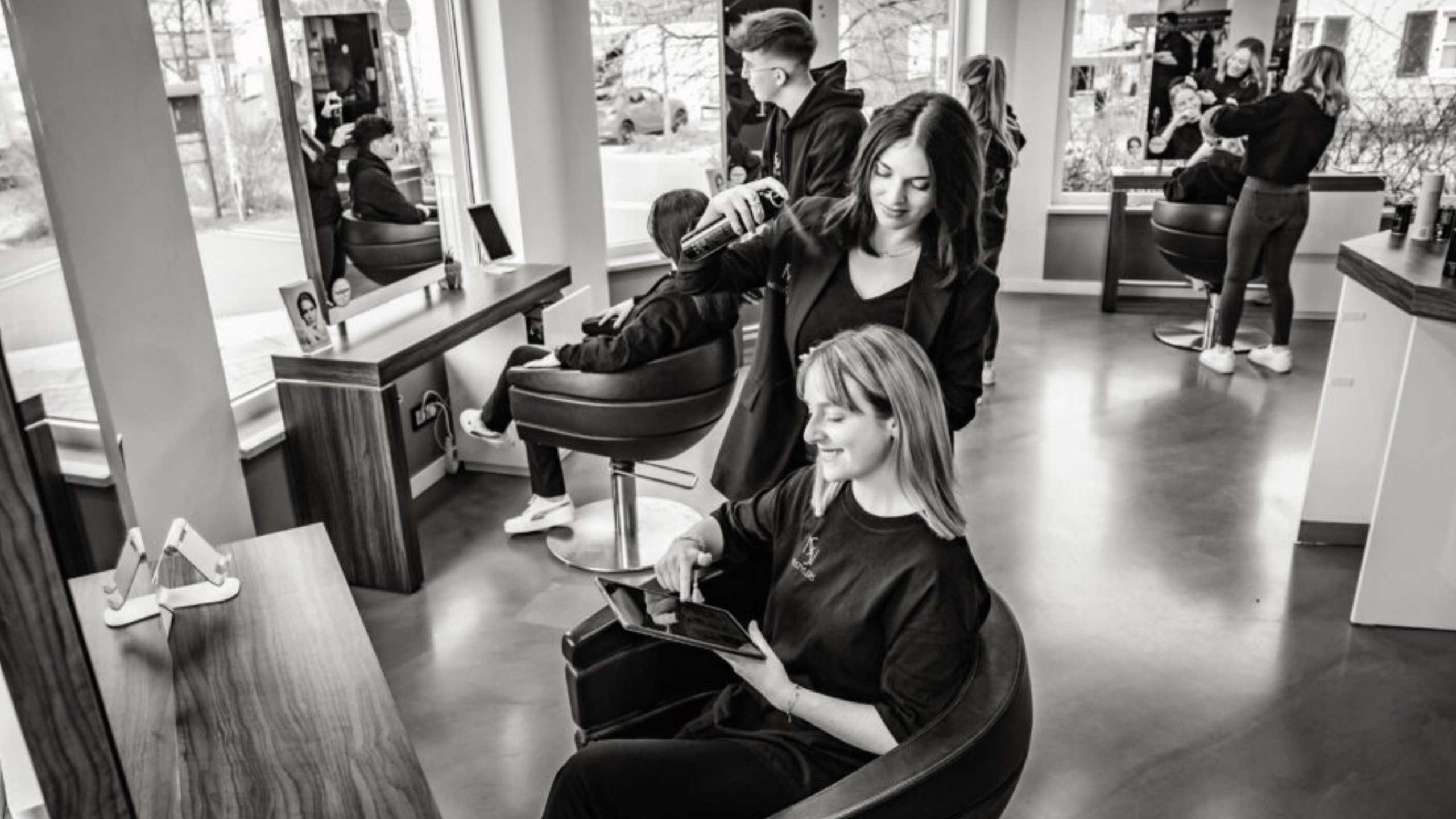 Ein belebter Friseursalon in Schwarz-Weiß. Friseure arbeiten an Kunden, die auf modernen Stühlen sitzen. Eine Frau lächelt, während sie auf einem Tablet liest. Große Fenster lassen Tageslicht ein.
