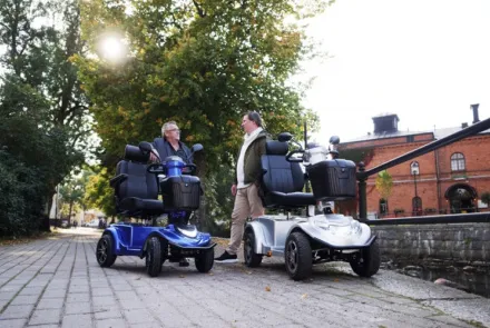Two adults stand and talk by their scooters on a paved path near a canal, with trees and a red brick building visible in the background on a sunny day.
