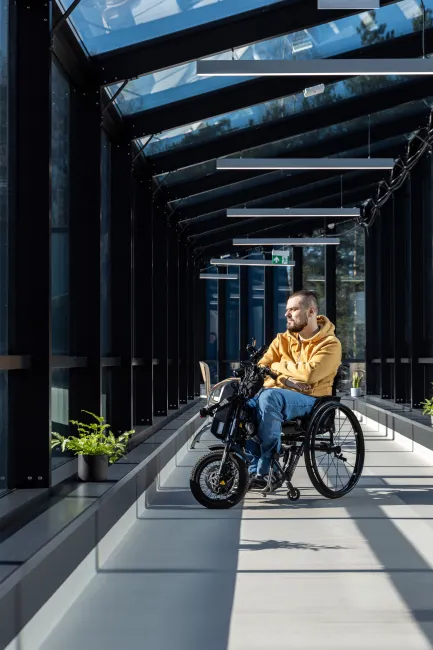 vīrietis riteņkrēslā apstājies gaitenī skatās ārā pa logu