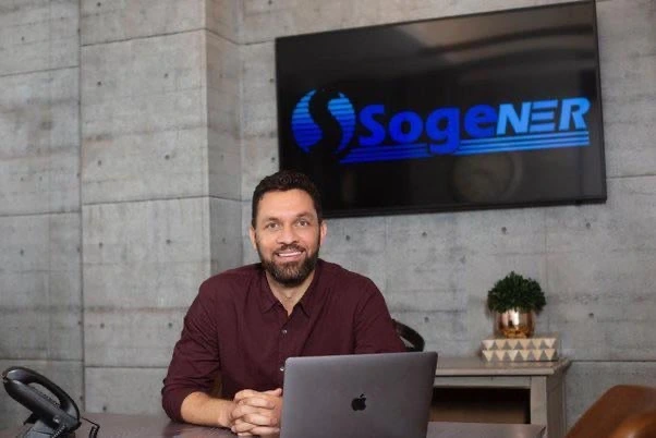 Man seated at a desk in front of a Sogener sign, smiling at the camera