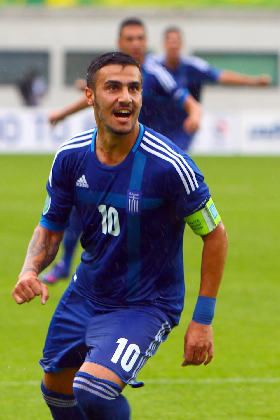 Giorgos Katidis in a Greek football match, wearing a blue Greece kit and captain's armband.