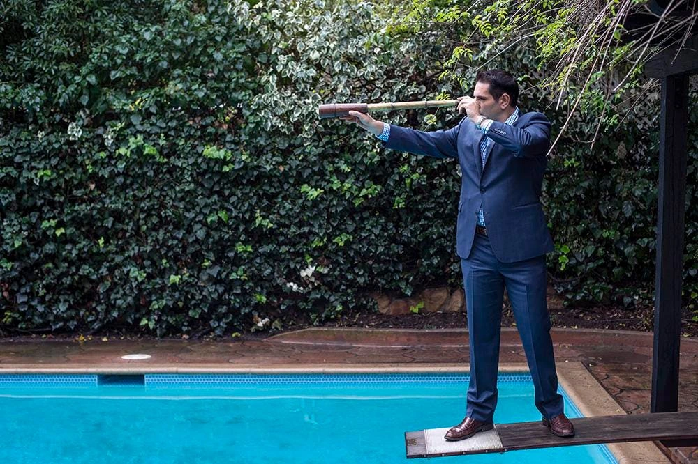 Demetri Argyropoulos in a blue suit standing by a pool with a long pipe or prop in an outdoor setting