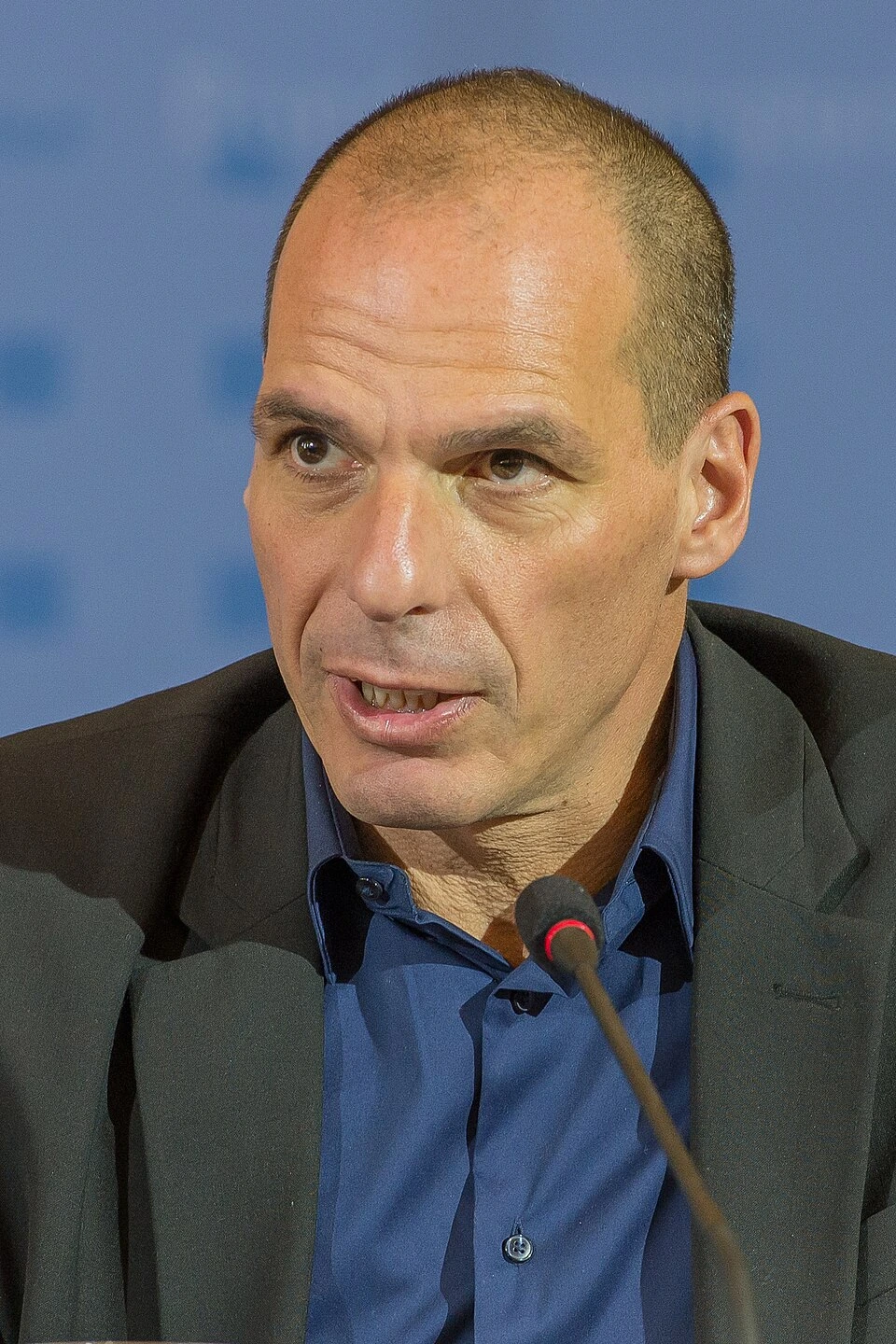 Yanis Varoufakis speaking at a podium during an event, wearing a dark jacket and blue shirt