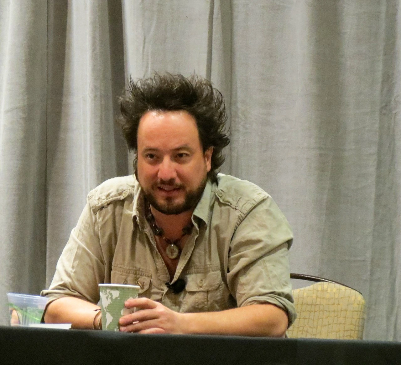 Giorgio A. Tsoukalos seated at a panel table, speaking in front of a curtain backdrop