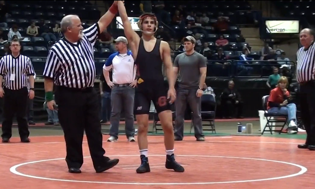 Georgio Poullas standing on a wrestling mat after a match, with a referee raising his arm in victory.