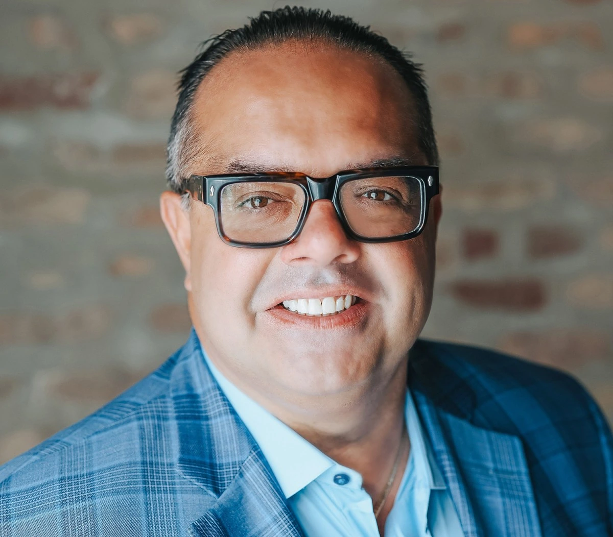 Portrait of Alex Pissios smiling in a blue suit jacket and glasses against a brick background.