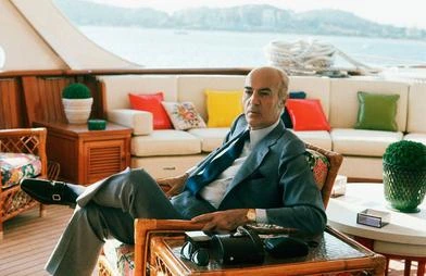 Basil Goulandris seated indoors on a yacht or luxury vessel, with the sea visible through the window behind him.