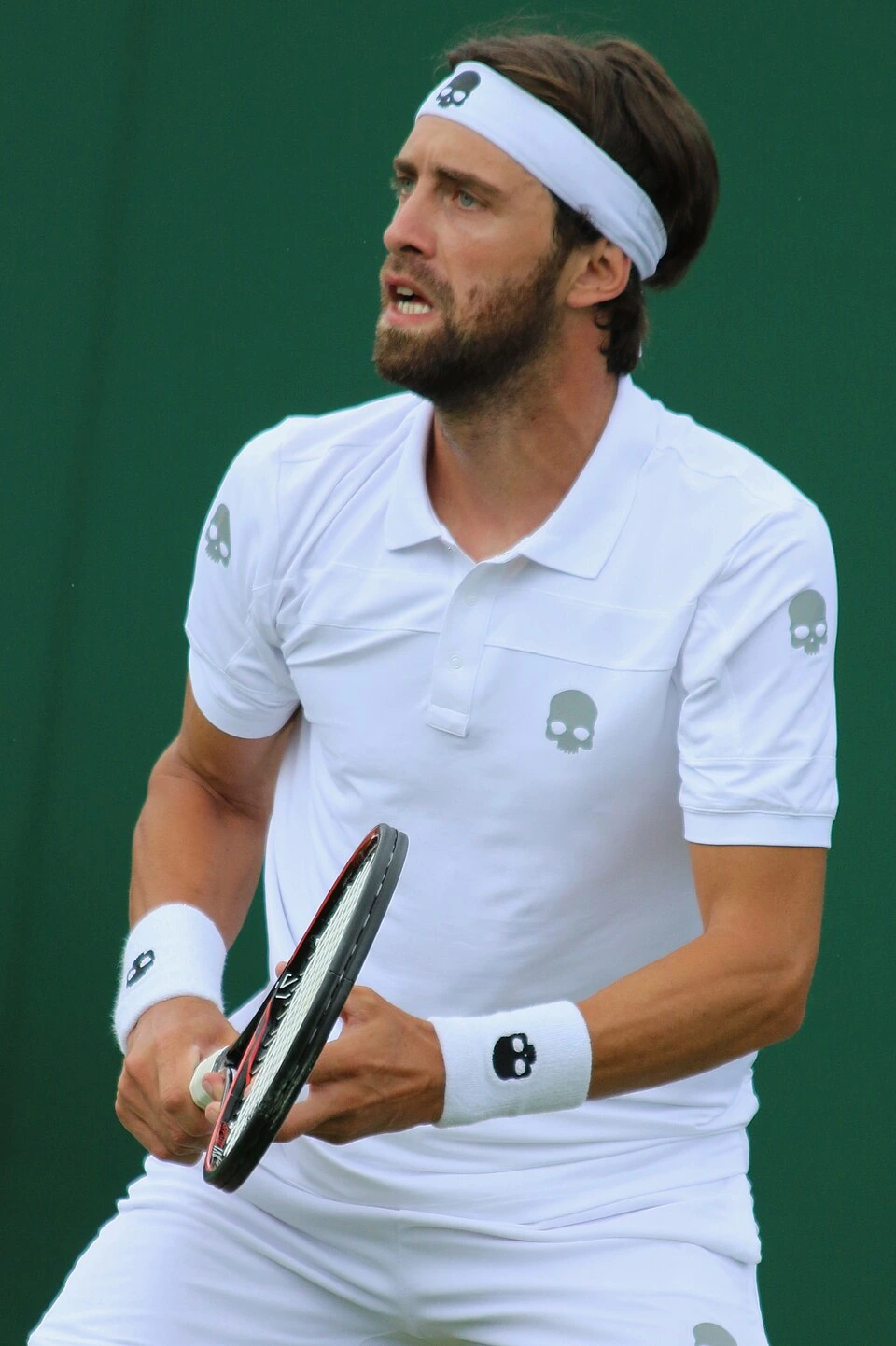 Nikoloz Basilashvili in a tennis match, wearing white kit and headband, holding a racket against a green court backgroun