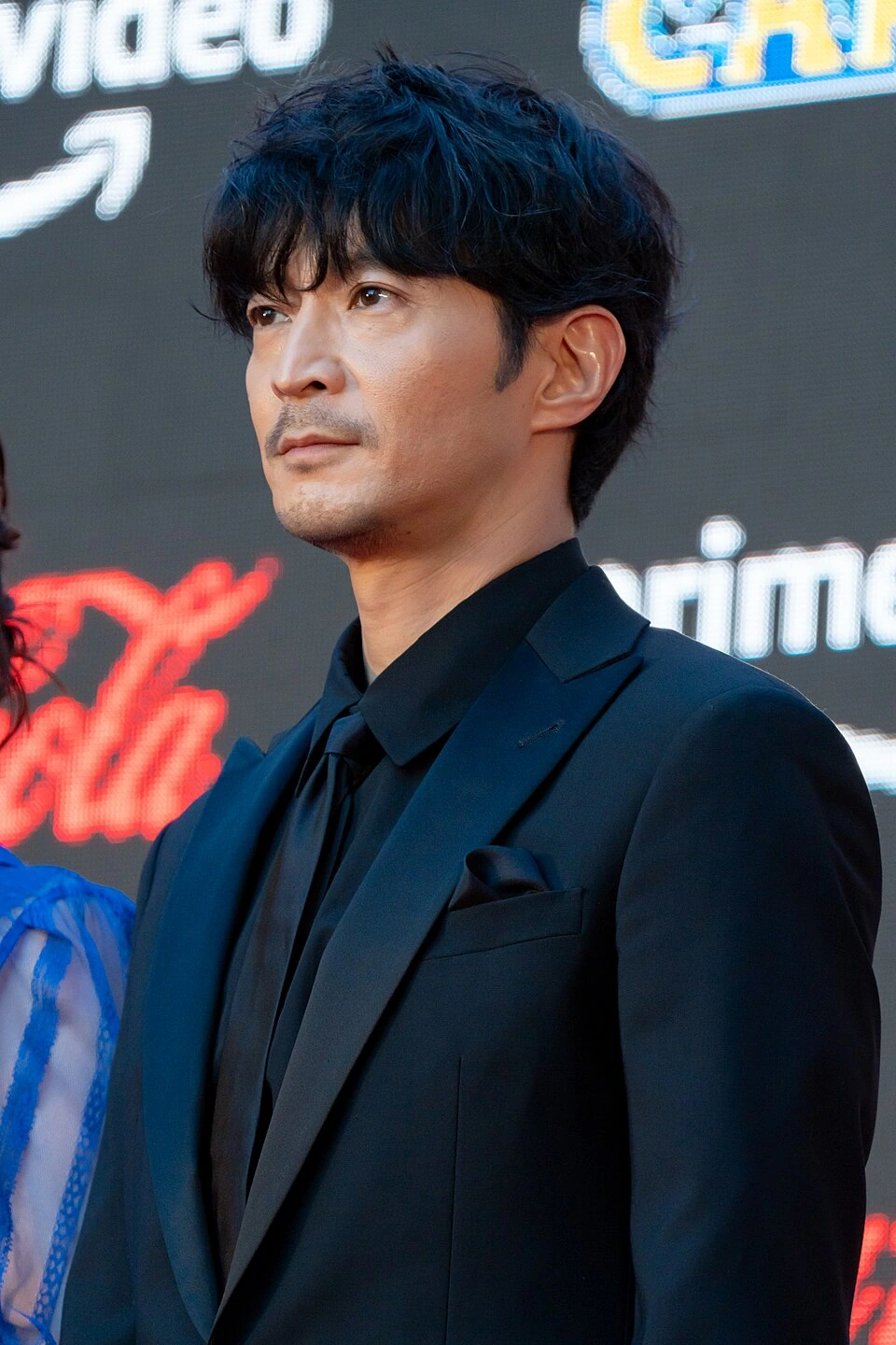 Kenjiro Tsuda in a black suit at a red carpet event, posed in front of a branded backdrop.