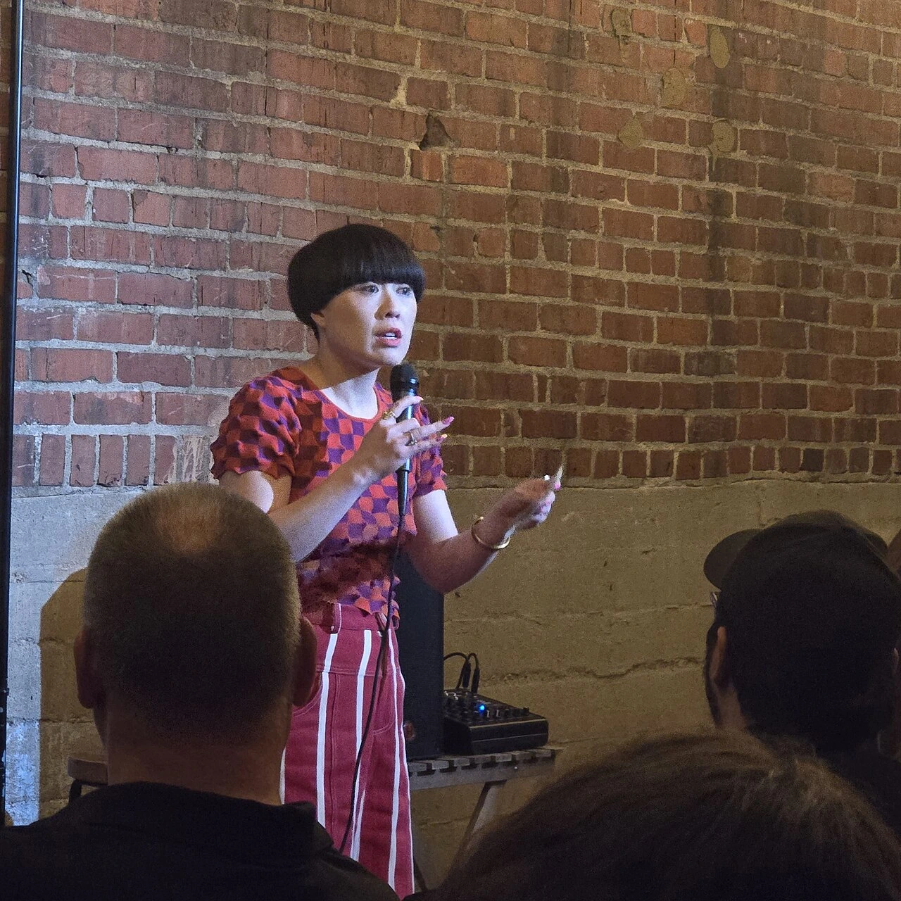 Atsuko Okatsuka performing stand-up in front of a brick wall, holding a microphone and speaking to an audience.