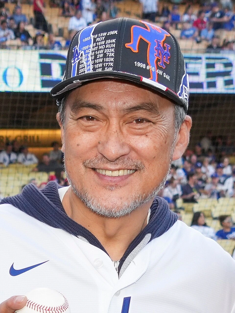 Ken Watanabe smiling at a baseball event, wearing a cap and white jersey