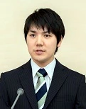 Kei Komuro speaking at a podium in a suit and tie