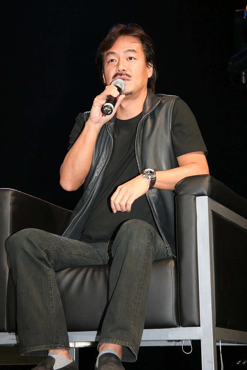 Hironobu Sakaguchi speaking at an event, seated on stage holding a microphone.