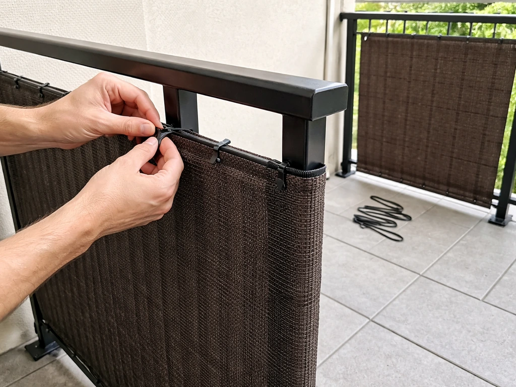 Hands zip-tying a privacy screen to a patio railing next to a second pre-made panel segment.