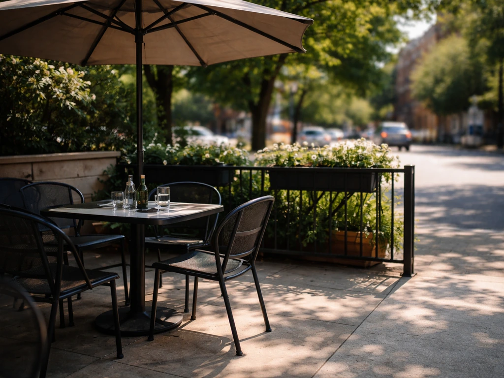 Outdoor patio with diners’ empty table under umbrella shade, direct sun spilling at the edge, faint traffic implied
