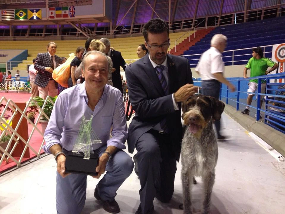 A man kneels beside a dog at an indoor event, holding a trophy while others stand in the background.