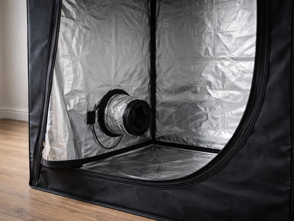 Close-up of an open grow tent showing reflective interior walls and intake/exhaust ports in a simple room.
