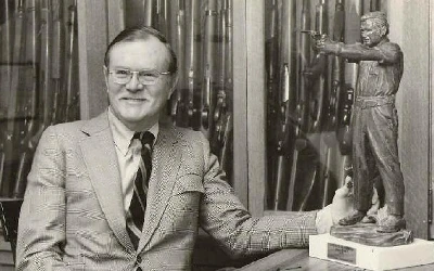 William B. Ruger seated beside a statue, in front of displayed firearms
