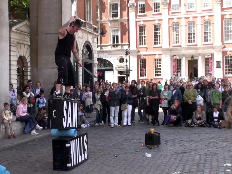 Sam Wills performing as Tape Face in a public square with a crowd watching