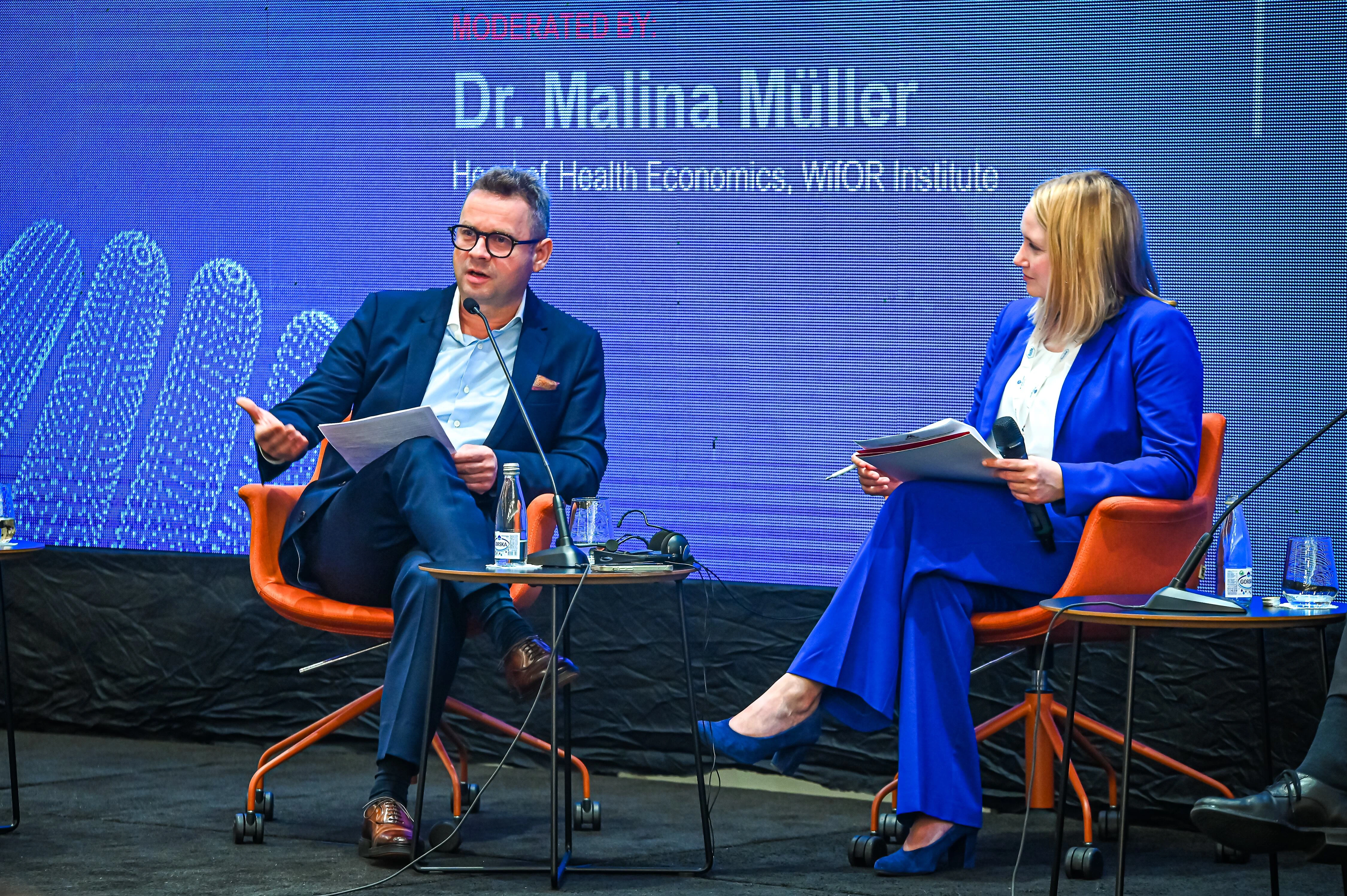 Živko Mukaetov seated on stage at a healthcare conference panel, speaking into a microphone beside another panelist