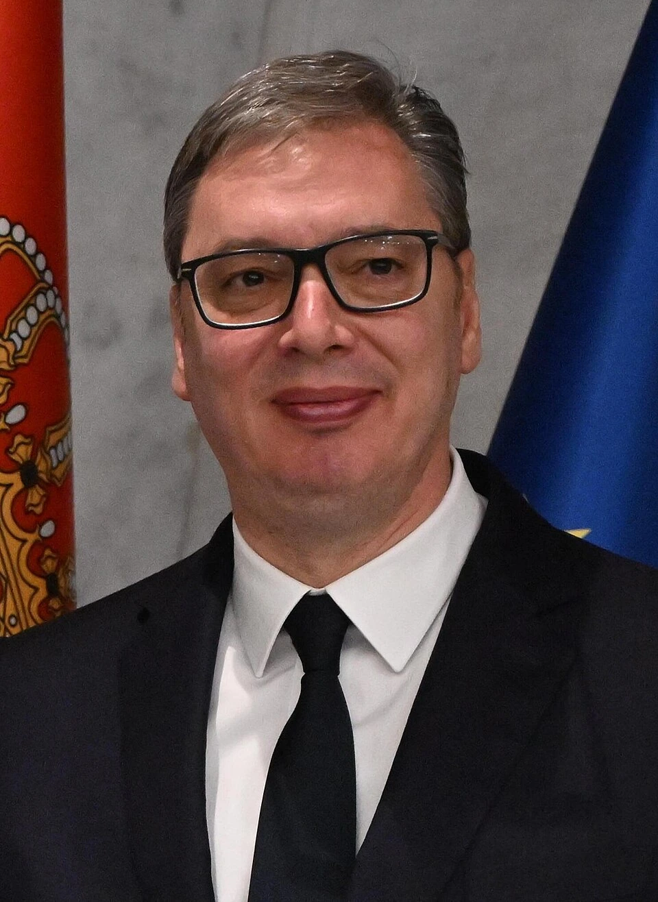 Aleksandar Vučić in a formal portrait wearing a black suit, white shirt, and black tie