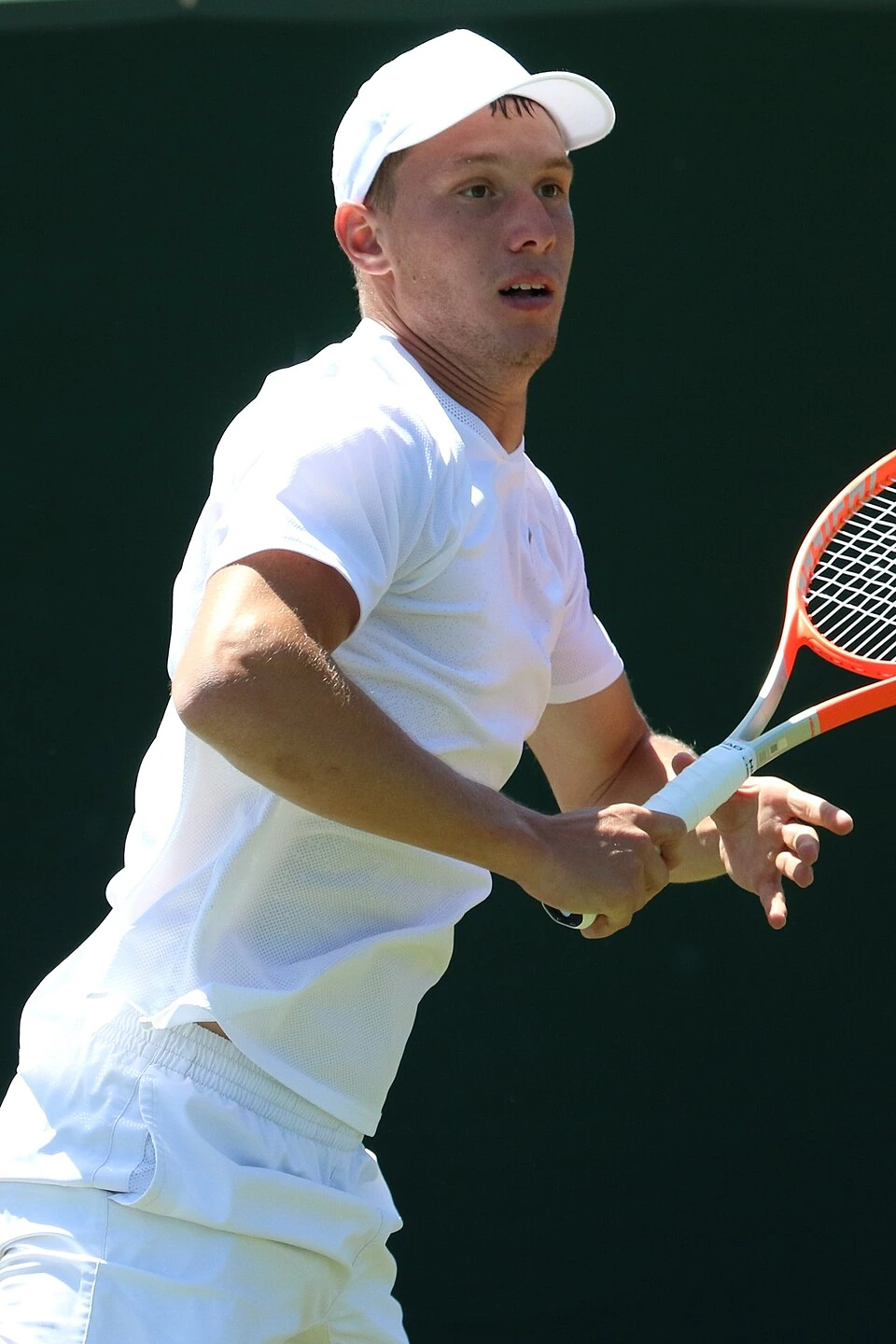 Filip Misolic playing tennis in a white outfit and cap, holding a racket on court
