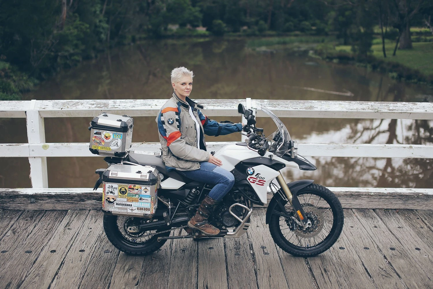 Kinga Tanajewska sitting on an adventure motorcycle on a wooden bridge with a river behind her
