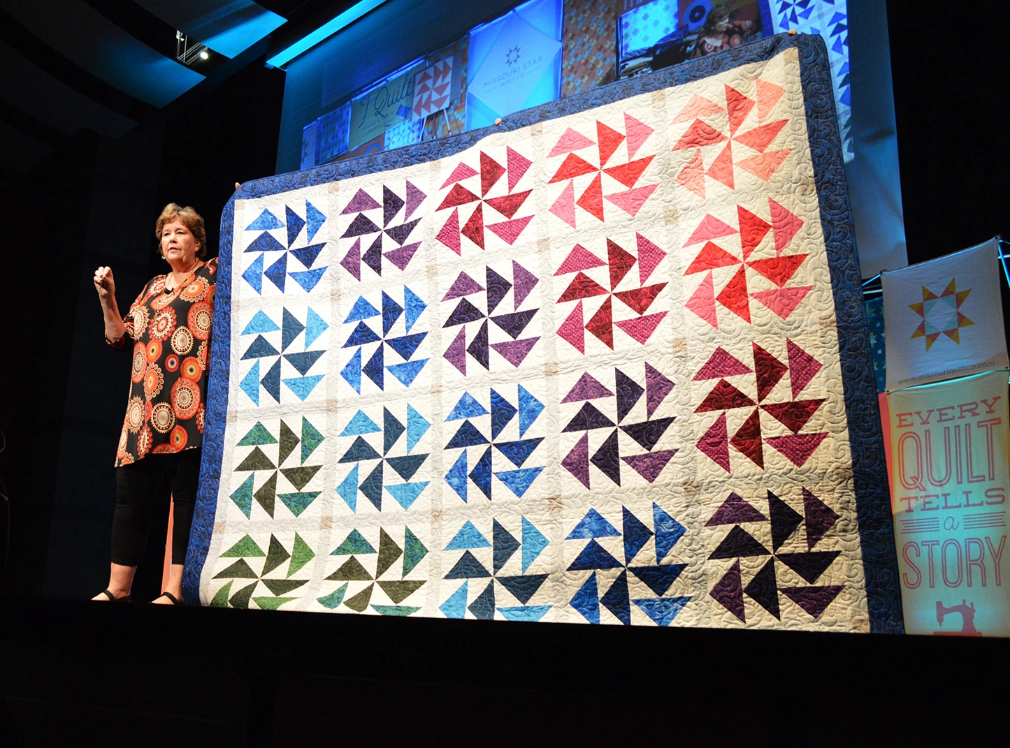 Jenny Doan speaking on stage while holding a colorful quilt at a Missouri Star Quilt Company event