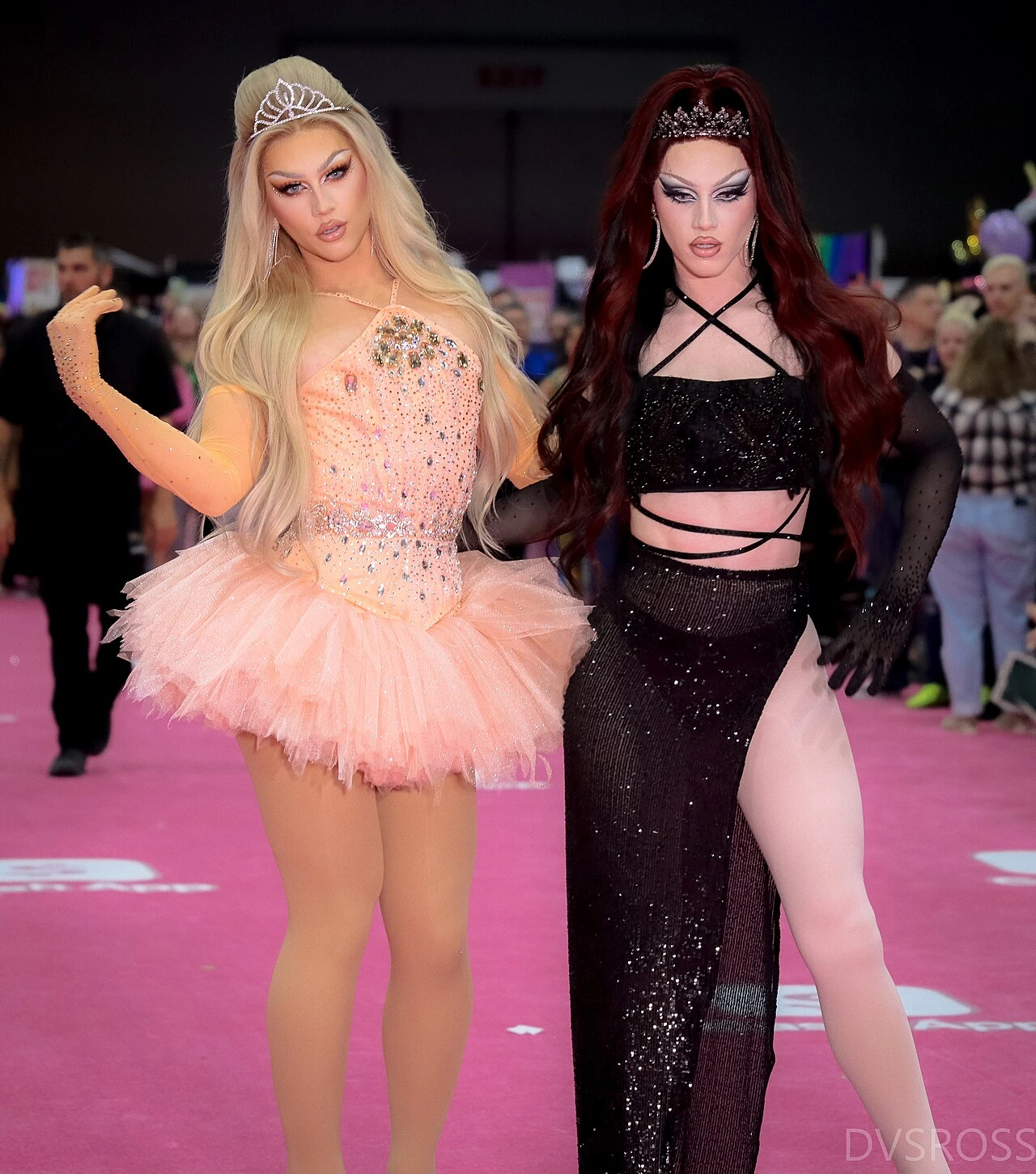 Two drag queens posed on a pink carpet at an event, wearing tiaras and glamorous outfits.