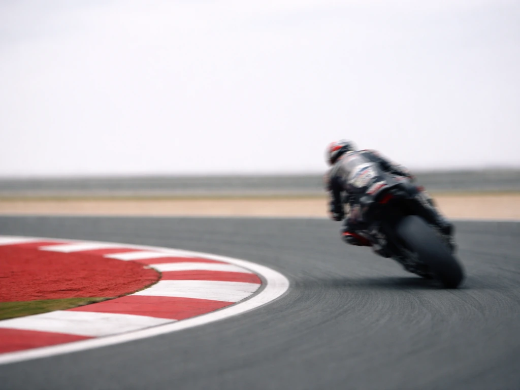 MotoGP-style race rider speeds past on a track, minimal scene symbolizing a MotoGP/WorldSBK era.